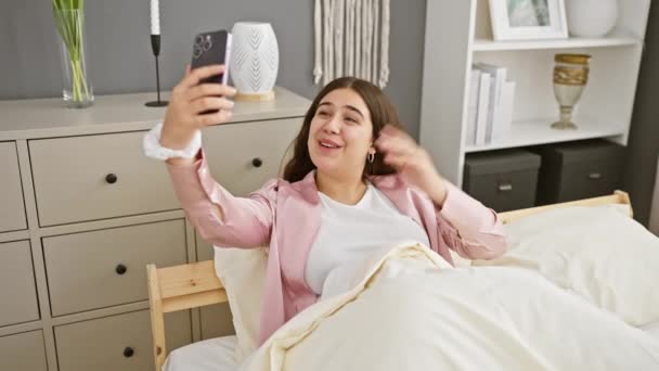 Lächelnde Frau macht Selfie im Schlafzimmer mit Smartphone und präsentiert lässigen Lebensstil und moderne Wohnkultur. - Filmmaterial, Video