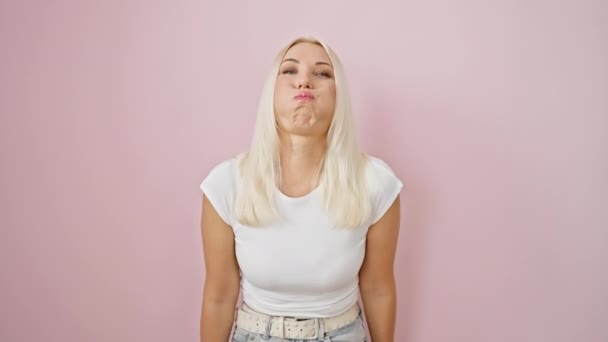 Смешная блондинка, стоящая на изолированном розовом фоне, пыхтя щеками в безумном смешном выражении лица, делая воздушное затяжение ртом - уверенно, но забавно! - Кадры, видео