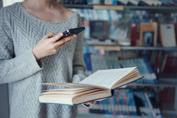 Молодая азиатская студентка колледжа с помощью смартфона сфотографировала страницу книги в школьной библиотеке. - Фото, изображение