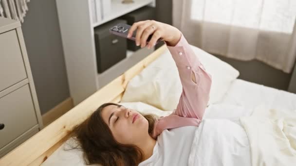 Eine junge Frau macht ein Selfie in ihrem hellen Schlafzimmer und zeigt ihr lässiges Leben zu Hause und ihre moderne Vernetzung. - Filmmaterial, Video