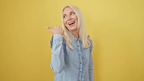 Щаслива молода блондинка, красиво впевнена, вказуючи великий палець вгору. весела жінка радісно демонструє позитивний жест рукою, стоячи ізольовано на жовтому тлі, одягнена в джинсову сорочку - Кадри, відео