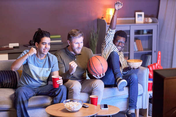 Různorodá skupina emocionálních sportovních fanoušků sledujících basketbal doma a jásání osvětlené modrým světlem - Fotografie, Obrázek