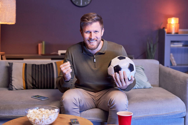 Porträt eines emotionalen Mannes, der zu Hause Sportspiele im Fernsehen verfolgt und den Ball im Blaulicht hält, Kopierraum - Foto, Bild