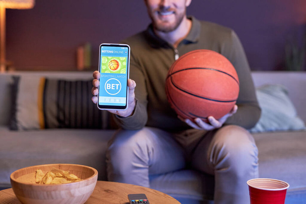 Close-up van de man met smartphone met online sportweddenschappen op het scherm en basketbal, kopieerruimte - Foto, afbeelding