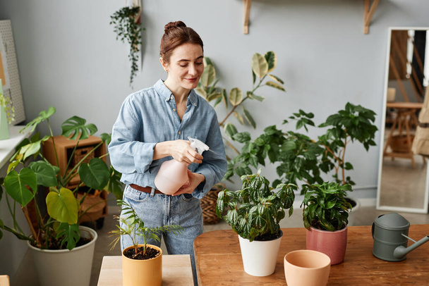 Retrato de una joven sonriente regando plantas en el interior y cuidando la vegetación del hogar, espacio para copiar - Foto, imagen