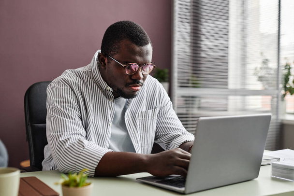 Portret czarnego mężczyzny w okularach i korzystającego z laptopa w swobodnym biurze - Zdjęcie, obraz