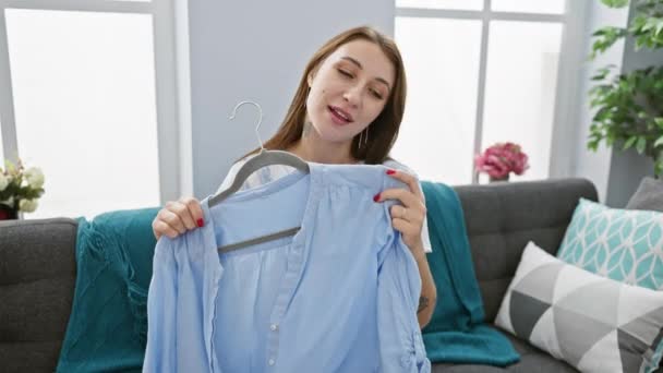 A young caucasian woman with brown hair holds a blue shirt on a hanger while sitting on a sofa in a well-lit living room. - Footage, Video