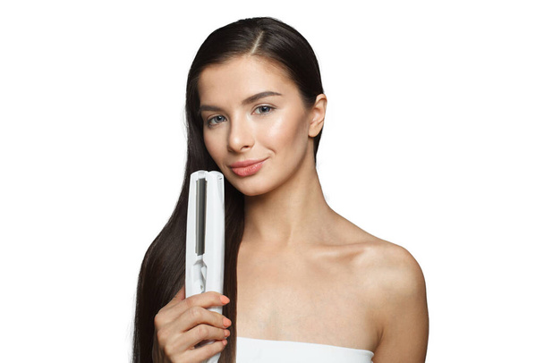 Славна модель жінка тримає залізо для волосся і випрямляє її здорове довге темне волосся, ізольоване на білому тлі. Концепція укладання волосся та волосся - Фото, зображення