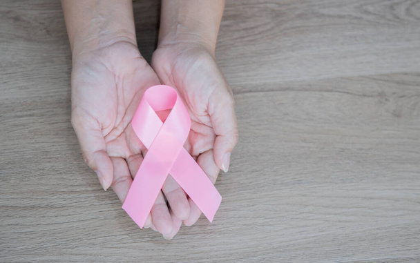 Χέρια με ροζ κορδέλα ως σύμβολο του αγώνα κατά του καρκίνου του μαστού σε ξύλινο φόντο. Έννοια της ευαισθητοποίησης σε θέματα υγείας, ιατρικής και καρκίνου του μαστού. - Φωτογραφία, εικόνα
