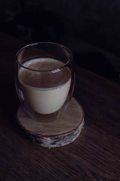Café cremoso sobre soporte de madera sobre fondo oscuro. Espuma de leche con espresso en vaso. Frappe en vidrio sobre mesa. Capuchino frío en taza. Latte cremoso batido. Bebida de aroma matutino. Bebidas lechosas. - Foto, Imagen