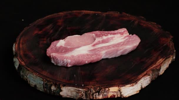 Un chef saltea y especia un trozo de carne de cerdo fresca cruda para freír (carne de cerdo) en una tabla de madera kraft. Primer plano sobre un fondo negro. Vista frontal - Imágenes, Vídeo