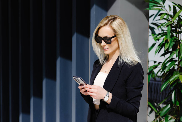 Jóvenes empresarias en traje negro usando teléfono inteligente aislado en la pared gris de fondo. Gerente bastante celebración de gadget móvil y mensaje de texto. - Foto, imagen