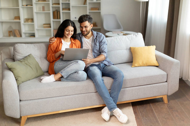 Planning vacation. Happy young indian couple searching tour online, using laptop computer, sitting on couch in cozy living room interior at home, free space - Photo, Image