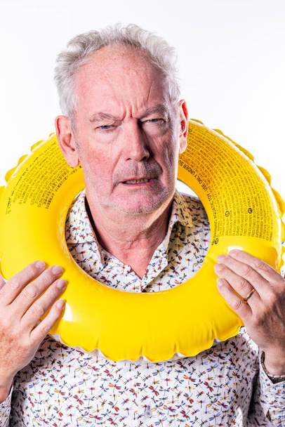 Dit beeld vangt een oudere man met een sceptische of onzekere uitdrukking terwijl hij een gele zwemring om zijn nek houdt. Het contrast tussen het heldere, speelse object en zijn dubieuze uitdrukking - Foto, afbeelding
