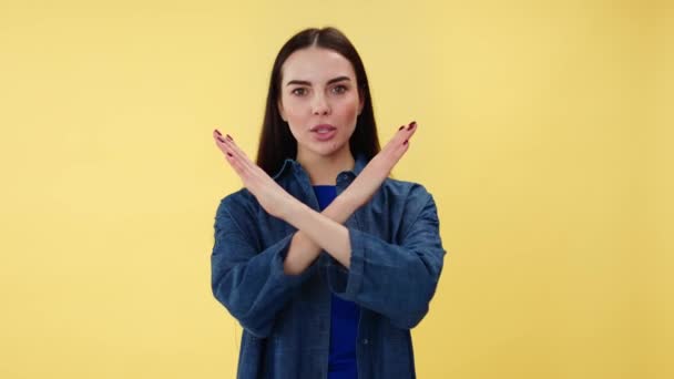Portret van een jonge vrouw met een serieuze gezichtsuitdrukking die handen kruist voor het tonen van stop gebaar. Verveelde brunette weigert iets met een teken van drooglegging. Geïsoleerd over gele studio achtergrond. - Video