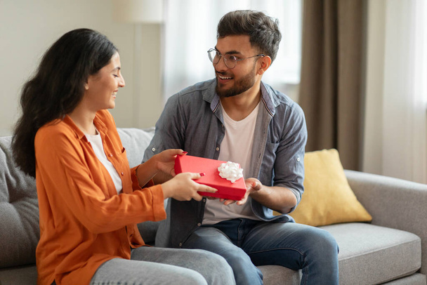 Happy indian spouses exchanging gifts while sitting on sofa at home, loving man giving wrapped present to his excited girlfriend, greeting each other - Photo, Image