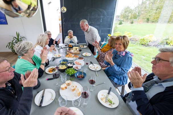 Ryhmä eläkeläisiä nauttii mukavan aterian yhdessä ruokapöydän ympärillä, joka on täynnä erilaisia ruokia ja viiniä. Mies seisoo, mahdollisesti nostaen maljan tai jakaen tarinan, kun häntä tervehditään - Valokuva, kuva