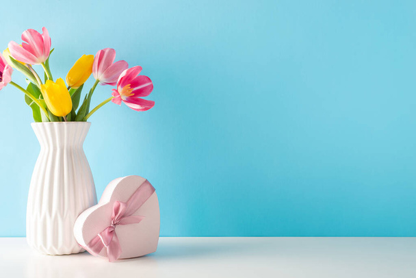 Muttertags- und 8. März-Thema: Beistelltisch mit herzförmigem Geschenk und einer Vase mit lebendigen Tulpen vor sanftem blauen Hintergrund, die Platz für Ihre Botschaft bietet - Foto, Bild