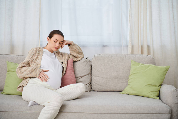 Een mooie zwangere vrouw zit comfortabel op een bank in een gezellige woonkamer, omgeven door warmte en anticipatie. - Foto, afbeelding