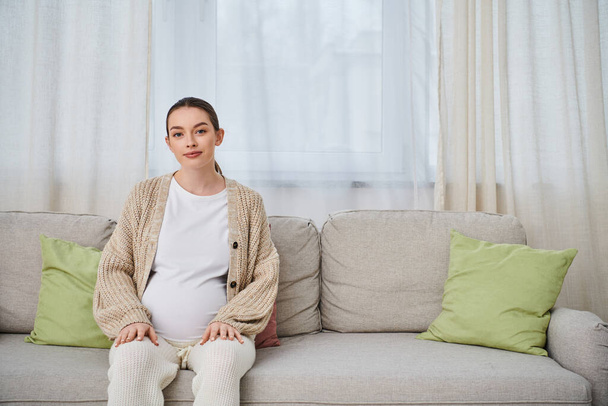 Una hermosa mujer embarazada se sienta en un sofá, su silueta iluminada por la luz suave de una ventana. - Foto, Imagen