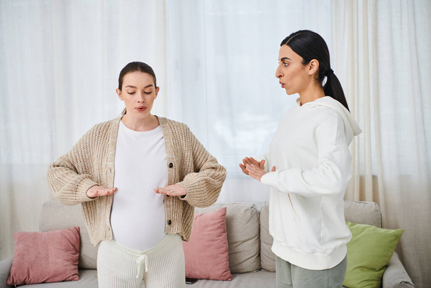 Eine schwangere Frau sitzt bei Elternkursen in einem gemütlich dekorierten Wohnzimmer neben ihrem Trainer auf einem Sofa. - Foto, Bild