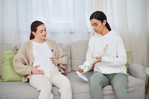 Μια έγκυος γυναίκα και ο εκπαιδευτής της κάθονται σε έναν καναπέ, κοιτάζοντας προσεκτικά το ανατομικό μοντέλο, μαθήματα γονέων. - Φωτογραφία, εικόνα