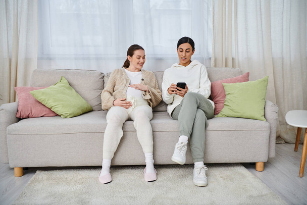 Egy férfi és egy terhes nő ül egy kanapén, lefoglalva egy mobiltelefon képernyőjén, valószínűleg megosztva egy pillanatnyi kapcsolatot. - Fotó, kép