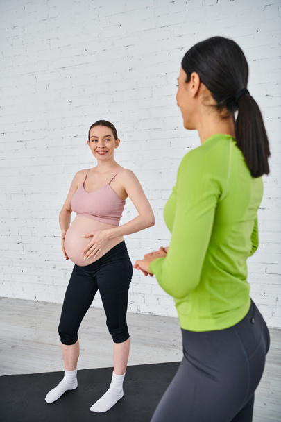 Μια έγκυος γυναίκα στέκεται με αυτοπεποίθηση μπροστά από ένα λευκό τούβλο στον τοίχο με τον προπονητή της κατά τη διάρκεια ενός μαθήματος γονέων. - Φωτογραφία, εικόνα