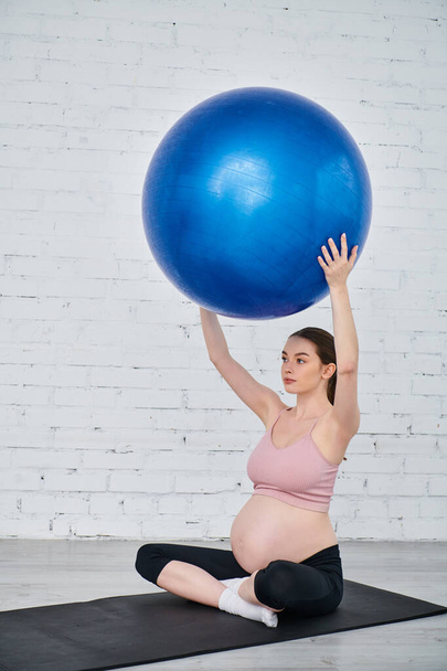 Těhotná žena v józe pózuje na podložce, drží modrý míč během prenatálního cvičení. - Fotografie, Obrázek