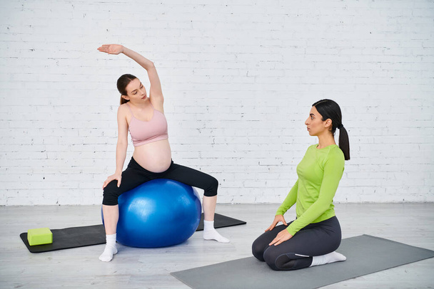 Μια έγκυος γυναίκα βρίσκει ισορροπία και δύναμη καθώς κάθεται στην κορυφή μιας μπλε μπάλας γυμναστικής κατά τη διάρκεια μαθημάτων γονέων με τον προπονητή της.. - Φωτογραφία, εικόνα