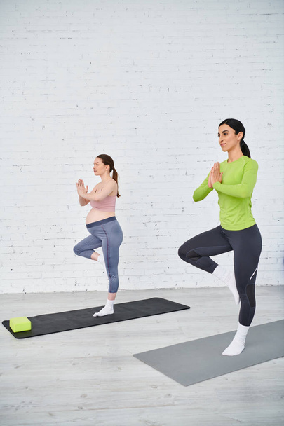 Dos mujeres, una embarazada, posan con gracia en posiciones de yoga, encontrando paz y fuerza frente a una pared de ladrillo blanco. - Foto, imagen