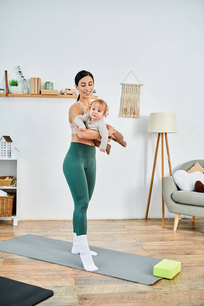 Egy fiatal anya áll a jóga szőnyegen, gyengéden fogja a babáját, egy edző vezetésével a szülői tanfolyamokon.. - Fotó, kép