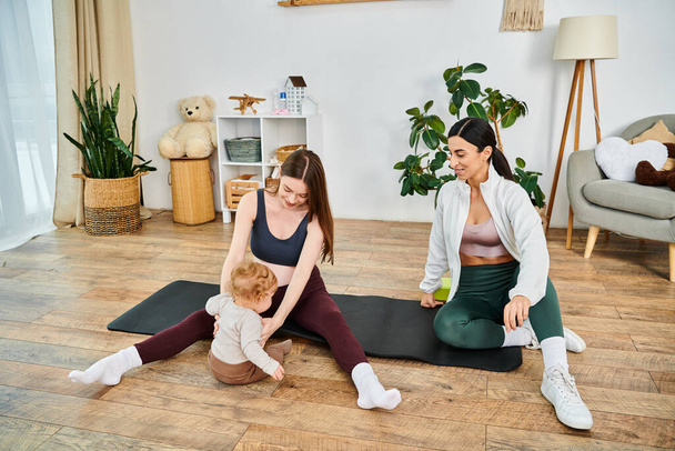 Két nő, egy derűs, fiatal anya és az edzője, egy csecsemőt vezetnek a jóga szőnyegen egy békés otthoni környezetben.. - Fotó, kép