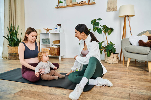 Młoda matka siedzi na macie do jogi ze swoim dzieckiem, prowadzona przez trenera na kursie rodzicielskim, wspierając chwilę spokoju i więzi.. - Zdjęcie, obraz