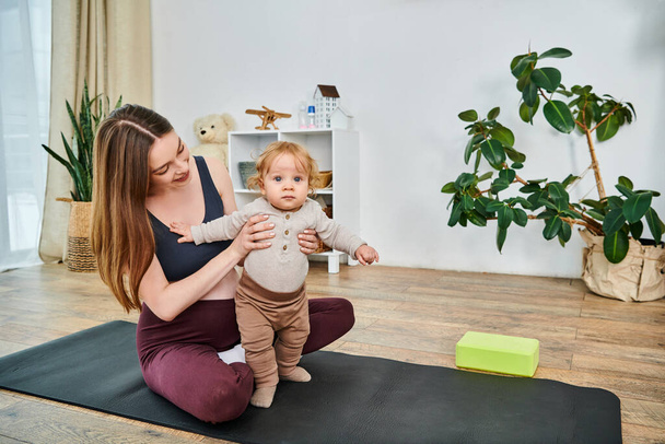 Eine junge Mutter sitzt auf einer Yogamatte und wiegt ihr Baby, angeleitet von ihrem Trainer in einem Moment der friedlichen Verbindung und Fürsorge. - Foto, Bild