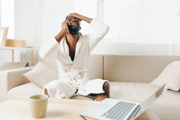 Homme afro-américain souriant travaillant sur ordinateur portable au bureau à domicile, parlant au téléphone dans le peignoir - Photo, image