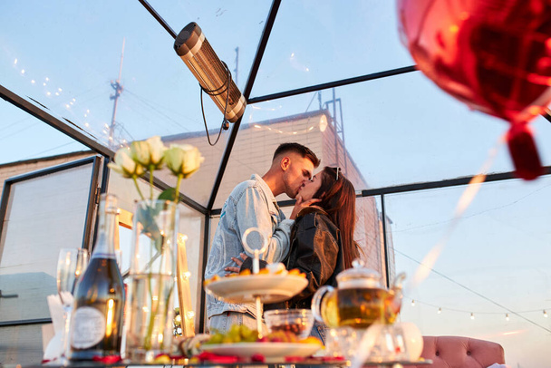 Um casal compartilha um momento terno em um telhado, suas silhuetas gravadas contra o céu da noite. O brilho da cidade melhora a atmosfera romântica. - Foto, Imagem