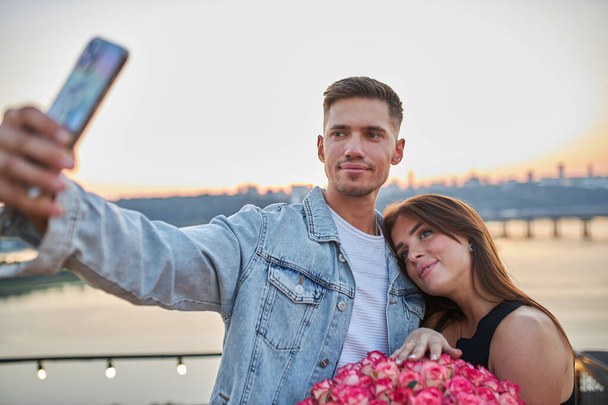 Una joven pareja con un enorme ramo de rosas se toma una selfie en una azotea, capturando el paisaje urbano al atardecer como telón de fondo. - Foto, imagen