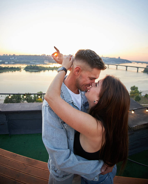 Nuori pari suutelee ja syleilee aikana romanttinen katolla päivämäärä, jossa kaupunkikuva taustan auringonlaskun. - Valokuva, kuva