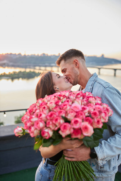 Ένα νεαρό ζευγάρι, σκαρφαλωμένο σε μια ταράτσα, μοιράζεται μια χαρούμενη στιγμή καθώς ο άνθρωπος χαρίζει ένα τεράστιο μπουκέτο τριαντάφυλλα στο φόντο ενός ορίζοντα της πόλης το ηλιοβασίλεμα.. - Φωτογραφία, εικόνα