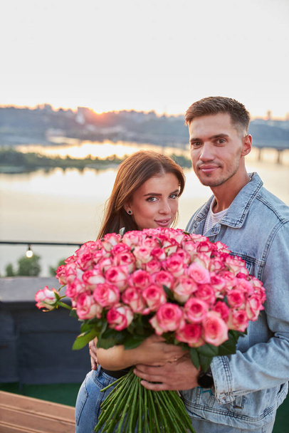 Un jeune couple, perché sur un toit, partage un moment heureux alors que l'homme offre un énorme bouquet de roses sur fond d'horizon urbain au coucher du soleil. - Photo, image