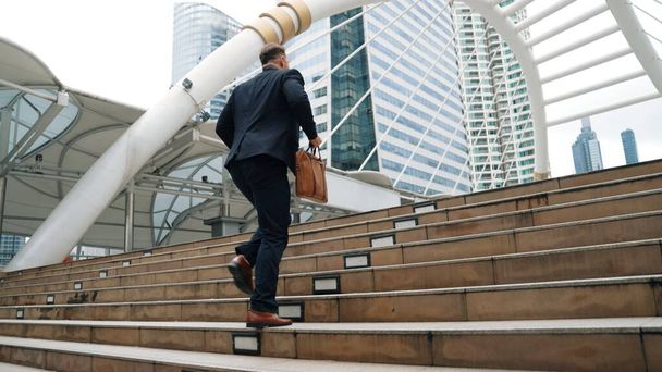 Έξυπνος καυκάσιος επιχειρηματίας ανεβαίνει τις σκάλες περιτριγυρισμένος από αστική θέα. Πλευρική άποψη του φιλόδοξου διαχειριστή του έργου εύρεση προκλητική δουλειά ή επενδυτική ευκαιρία στην πόλη με σύγχρονο κτίριο. Ουρμπάνος. - Φωτογραφία, εικόνα