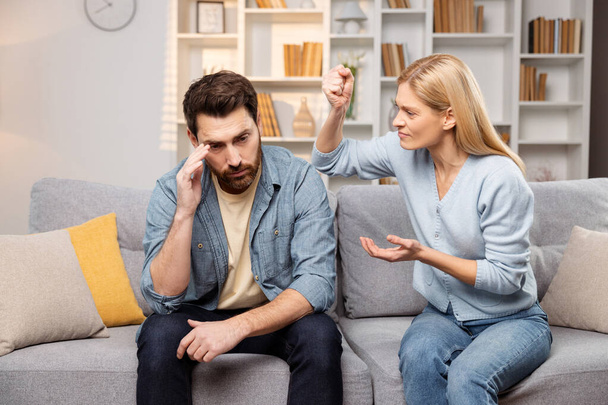 Érzelmi segély a küzdő családoknak, akiket férfiként és nőként ábrázolnak, akik hevesen vitatkoznak a kanapén, miközben a férj szorongatja a fejét. - Fotó, kép