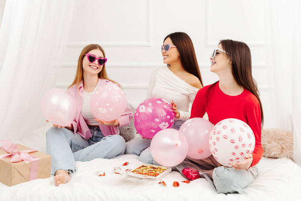 Glückliche schöne multiethnische Frauen mit stylischer Sonnenbrille sitzen auf dem Bett und schauen einander an. Junge attraktive Models posieren für Fotos - Foto, Bild