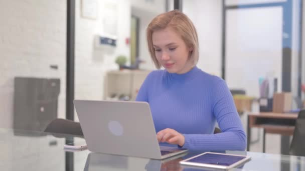 Nuori nainen hymyilee kamerassa työskennellessään kannettavan tietokoneen toimistossa - Materiaali, video