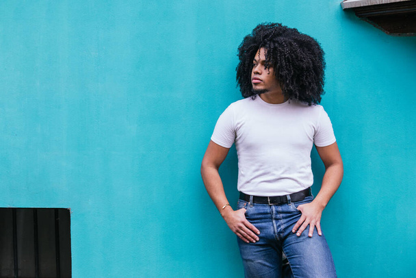 Contemplative Afro-Latino man standing with hands in pockets against a serene turquoise wall, exuding a relaxed urban vibe. - Photo, Image