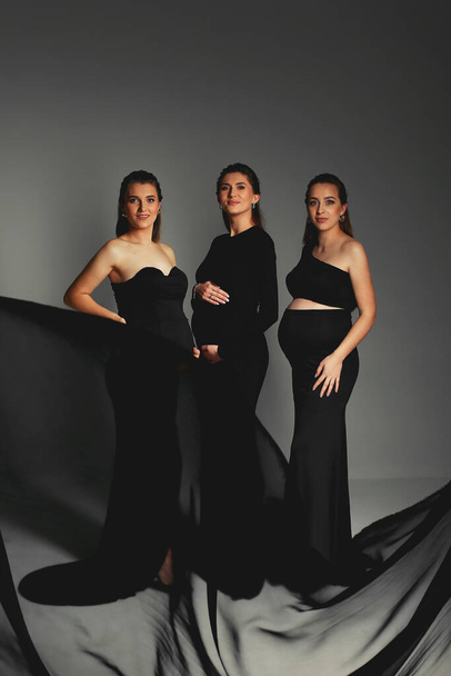  Група стильних вагітних жінок в чорних сукнях і позує в студії з чорним тканиною. Щасливі вагітні жінки в чорних сукнях з великими джинсами. Щасливе материнство  - Фото, зображення