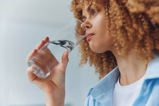 Κατσαρομάλλα γυναίκα απολαμβάνοντας ένα δροσιστικό ποτό νερού από ένα ποτήρι σε μια ηλιόλουστη μέρα - Φωτογραφία, εικόνα