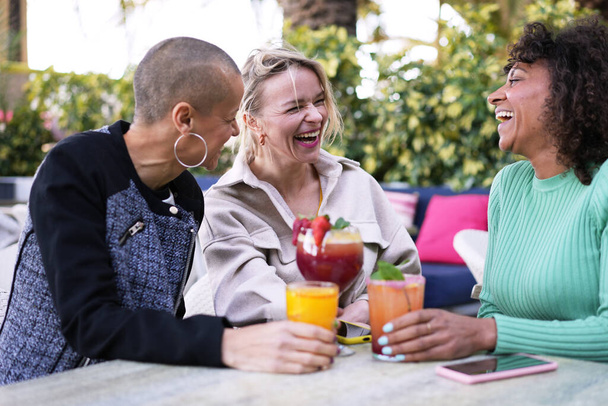 外で笑う成熟した多民族の女性の友人を集める陽気な集まり. パブのテラスで楽しいカクテルを飲む3人の成人女性の笑顔のグループ. 友情関係,アルコール飲料 - 写真・画像
