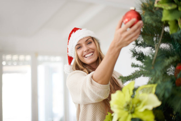 Γυναίκα, πορτρέτο και διακόσμηση δέντρων για τα Χριστούγεννα με χαμόγελο, διασκέδαση και ενθουσιασμό στο σπίτι. Εορταστική, Χριστούγεννα διακόσμηση και το κορίτσι με στολίδι, Σάντα καπέλο και ευτυχισμένη εκδήλωση διακοπών Δεκεμβρίου στο σαλόνι διαμέρισμα - Φωτογραφία, εικόνα